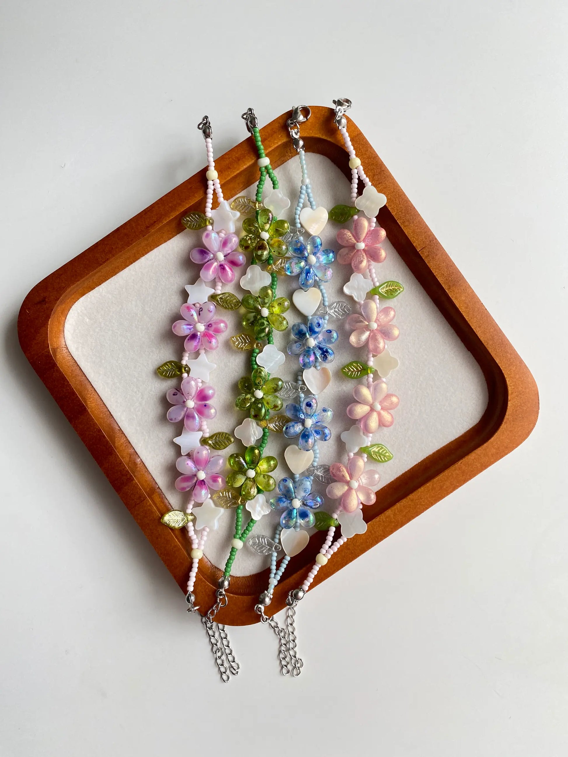 A collection of four colorful floral beaded bracelets featuring shell charms (heart, star, clover) displayed in a rustic wooden tray.