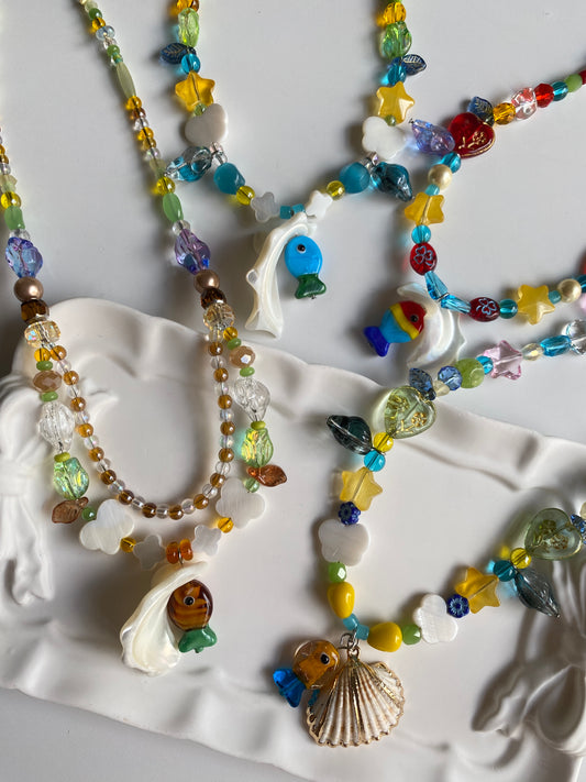Handcrafted ocean necklace collection photographed from above, featuring natural Triton shells, colorful fish glass charms and unique coastal-inspired beadwork.
