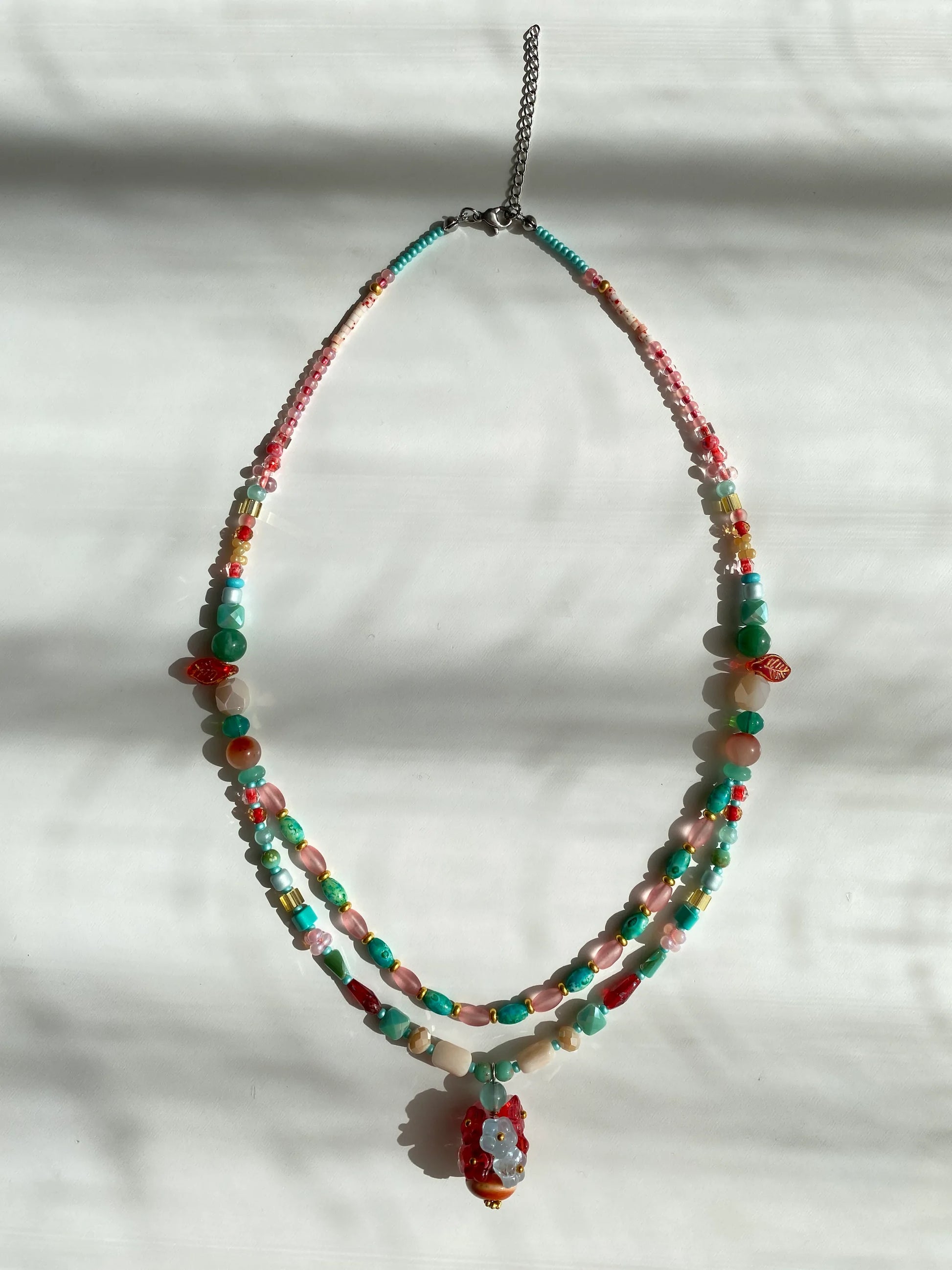 Detailed view of a handcrafted vibrant necklace featuring a crimson ceramic vase overflowing with 3D beaded flowers on a sunlit white desk.