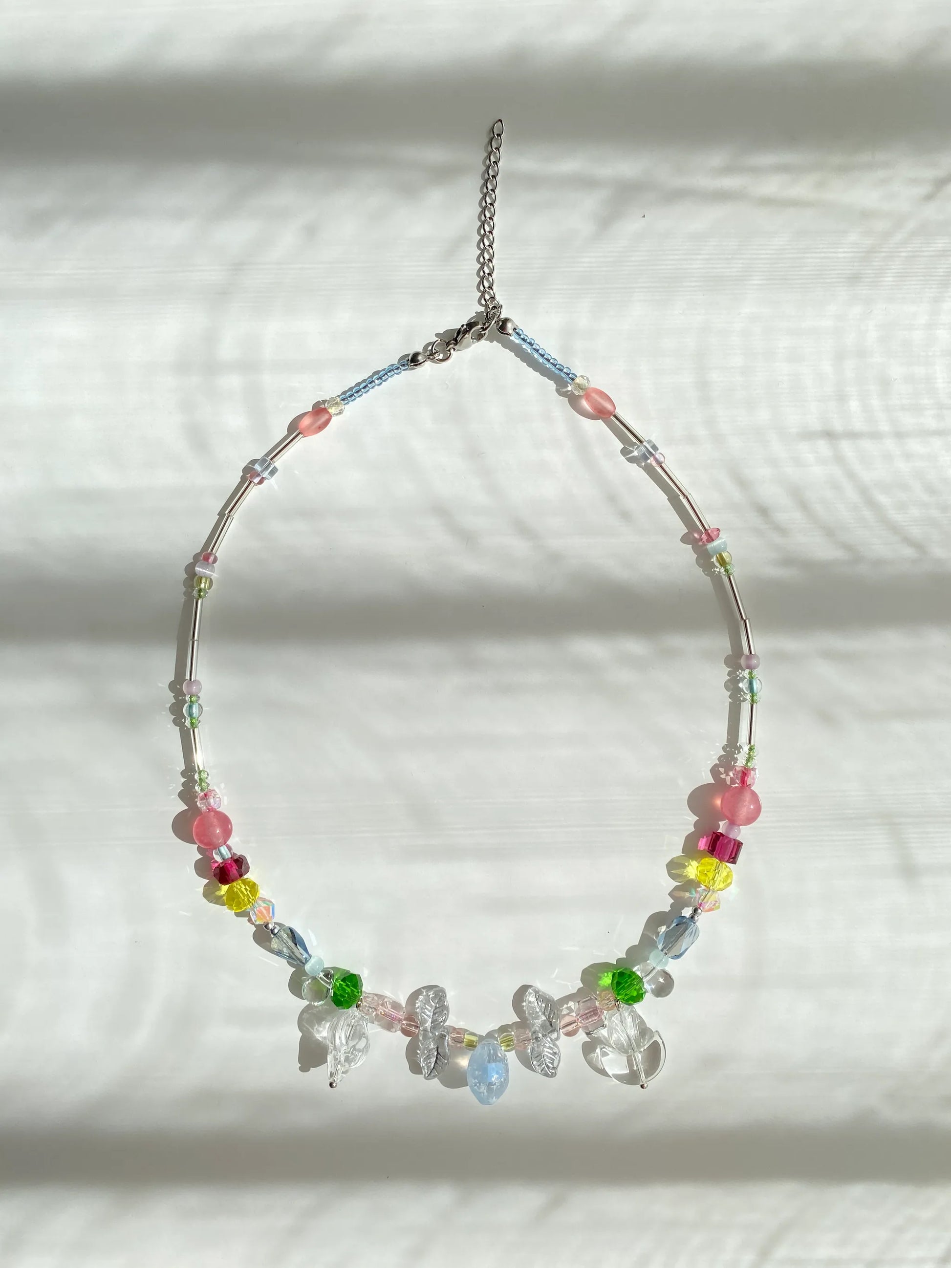 Handmade vibrant multi-color seed bead necklace featuring a glass lemon pendant, conch shell, and pink natural stones, flat lay on a sunlit table.