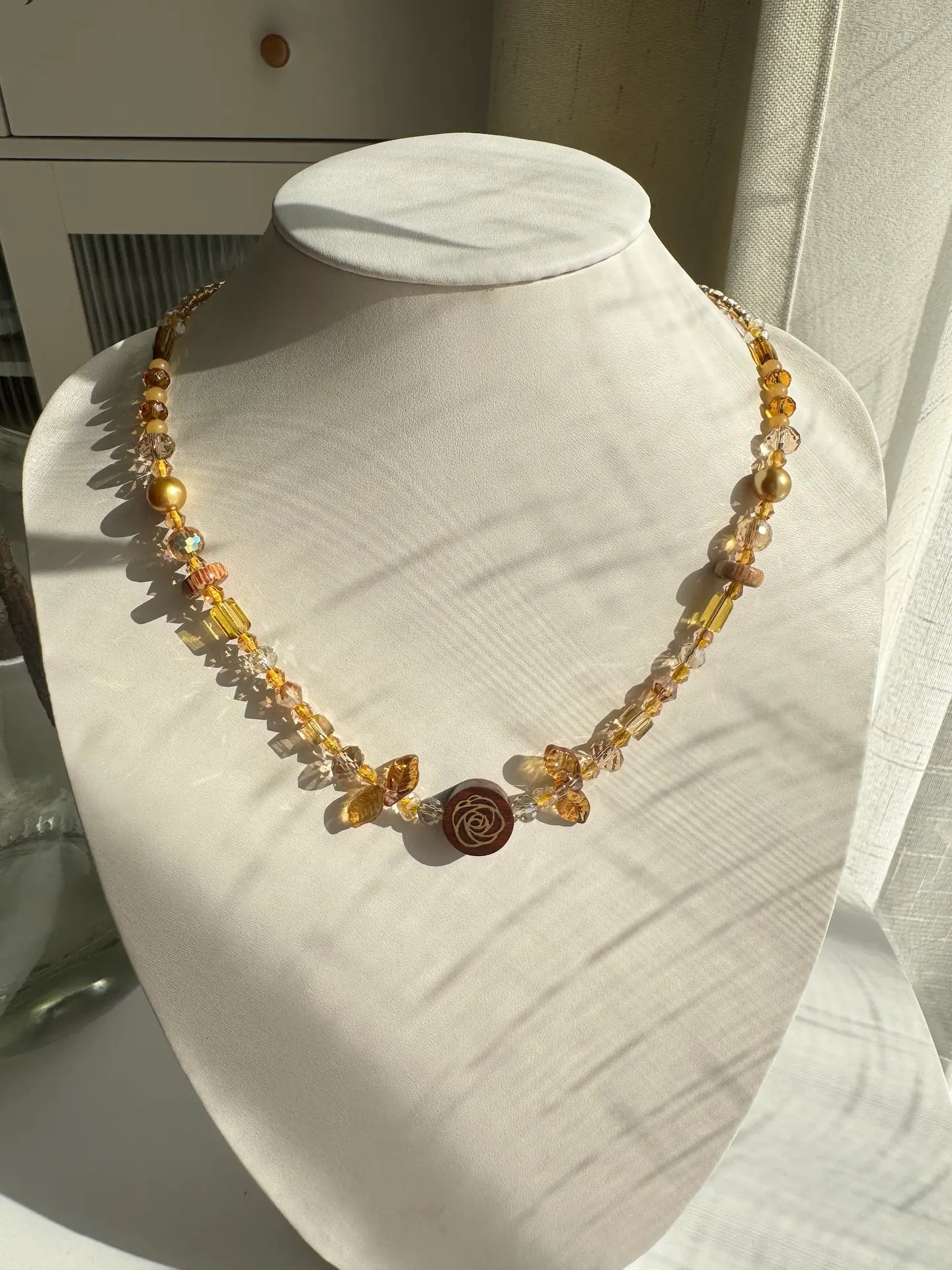 A 1-of-1 Maillard-style beaded necklace in khaki and brown tones featuring a central wooden rose pendant and leaf accents on a jewelry bust.