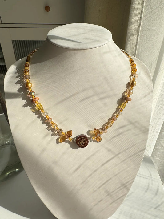 A 1-of-1 Maillard-style beaded necklace in khaki and brown tones featuring a central wooden rose pendant and leaf accents on a jewelry bust.