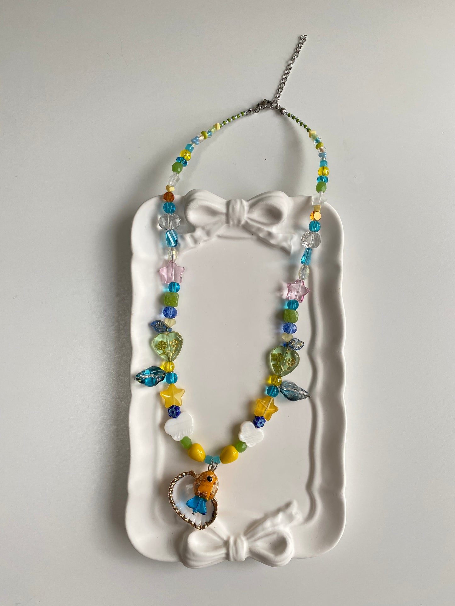 Orange and blue glass fish necklace displayed on a white surface with a fan shell pendant, butterfly shell charms and shimmering coastal glass beads.