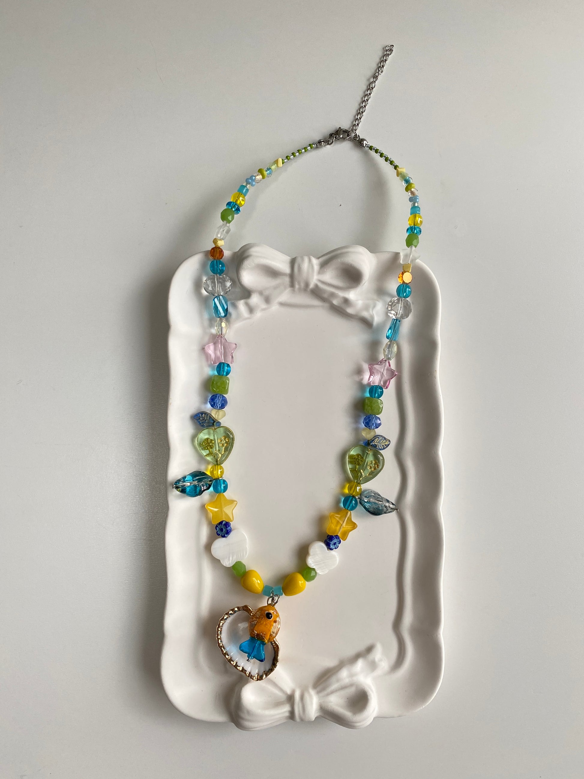 Orange and blue glass fish necklace displayed on a white surface with a fan shell pendant, butterfly shell charms and shimmering coastal glass beads.