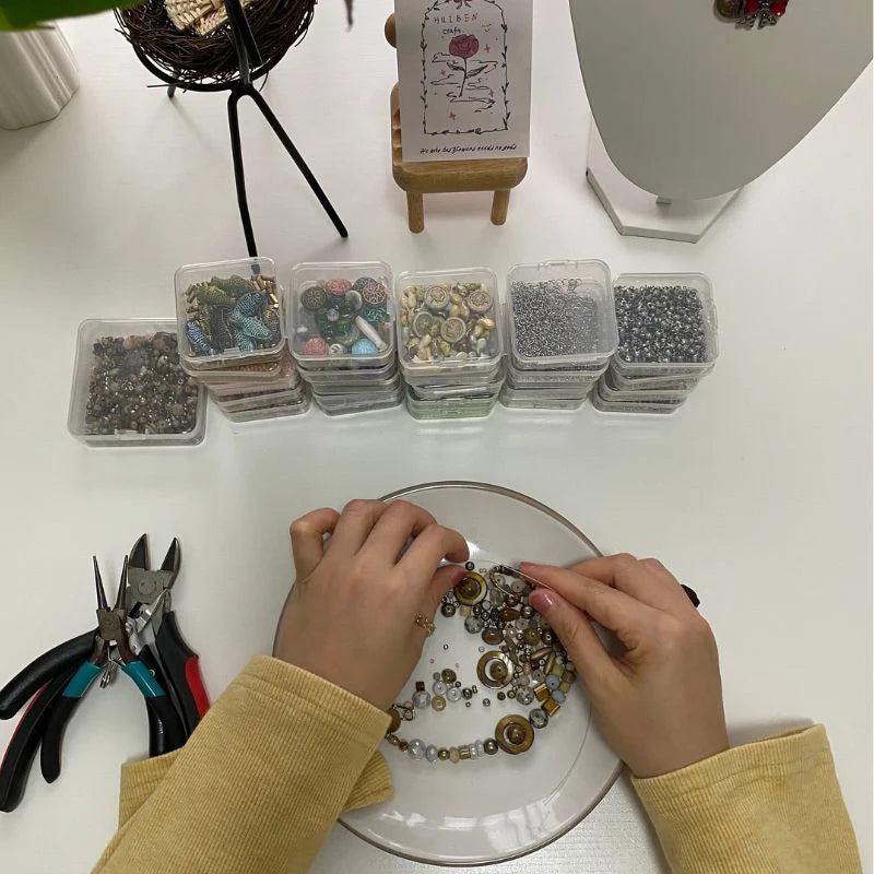 An overhead view of HuibenCraft founder crafting a handmade necklace at a white workspace, featuring pearl beads, jewelry pliers, and organized bead boxes under natural light.