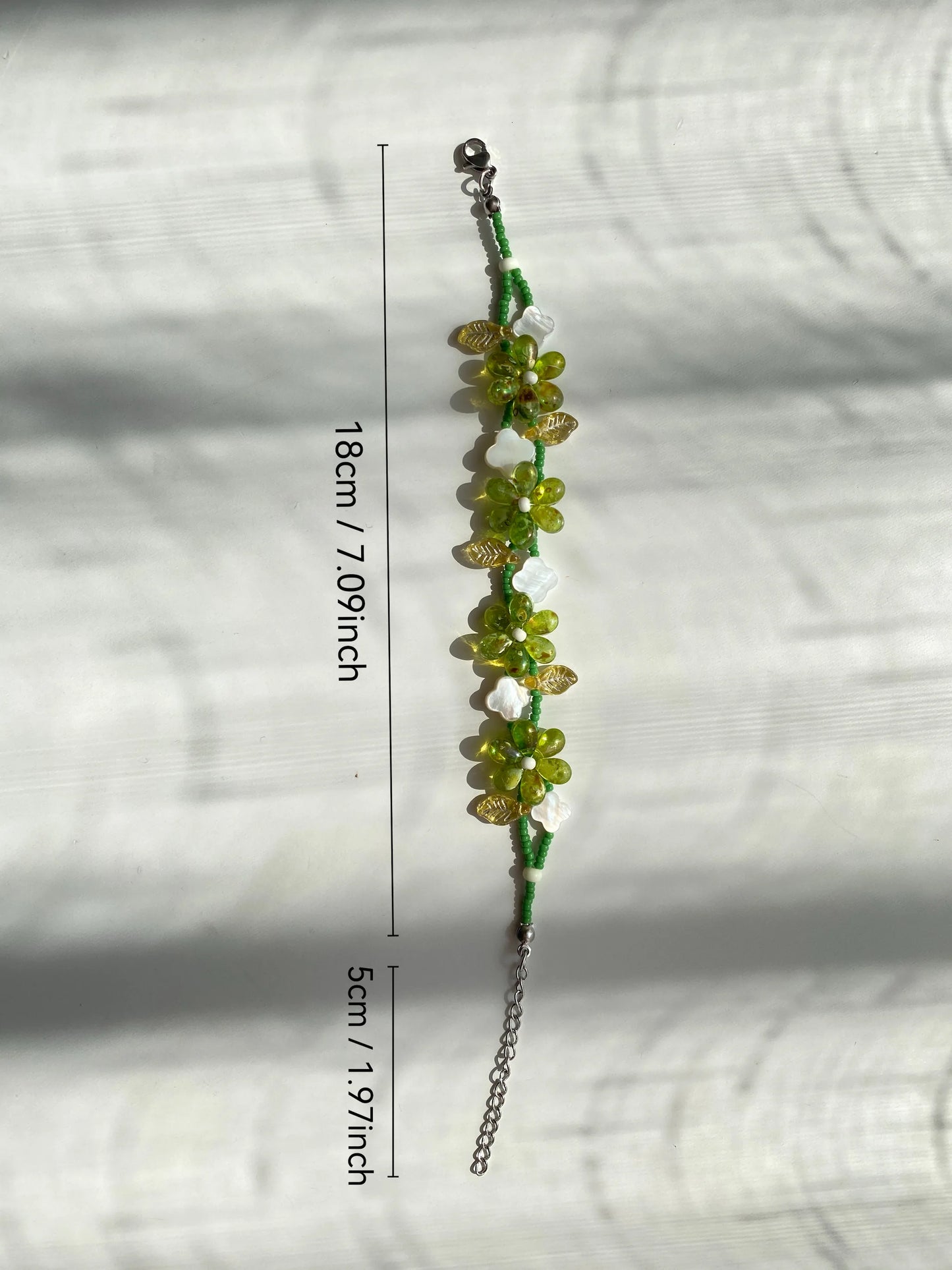 Unique 1-of-1 green floral beaded bracelet with a clover shell and leaf beads, artisanal jewelry on a sunlit white desk.