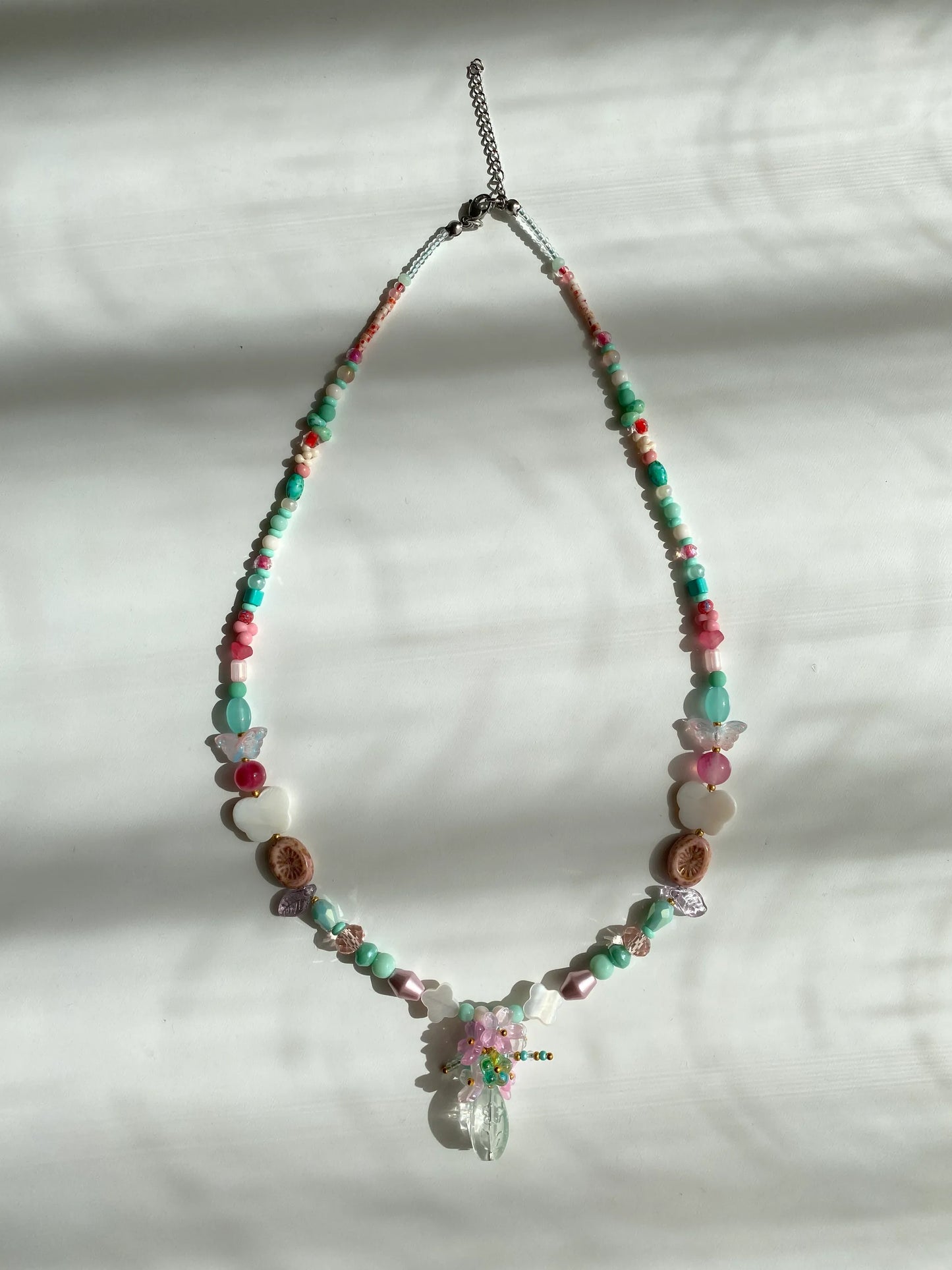 Detailed view of a unique pink and sky blue necklace with a beaded flower bouquet in a vase, featuring shell accents and kiwi glass beads on a sunlit white desk.