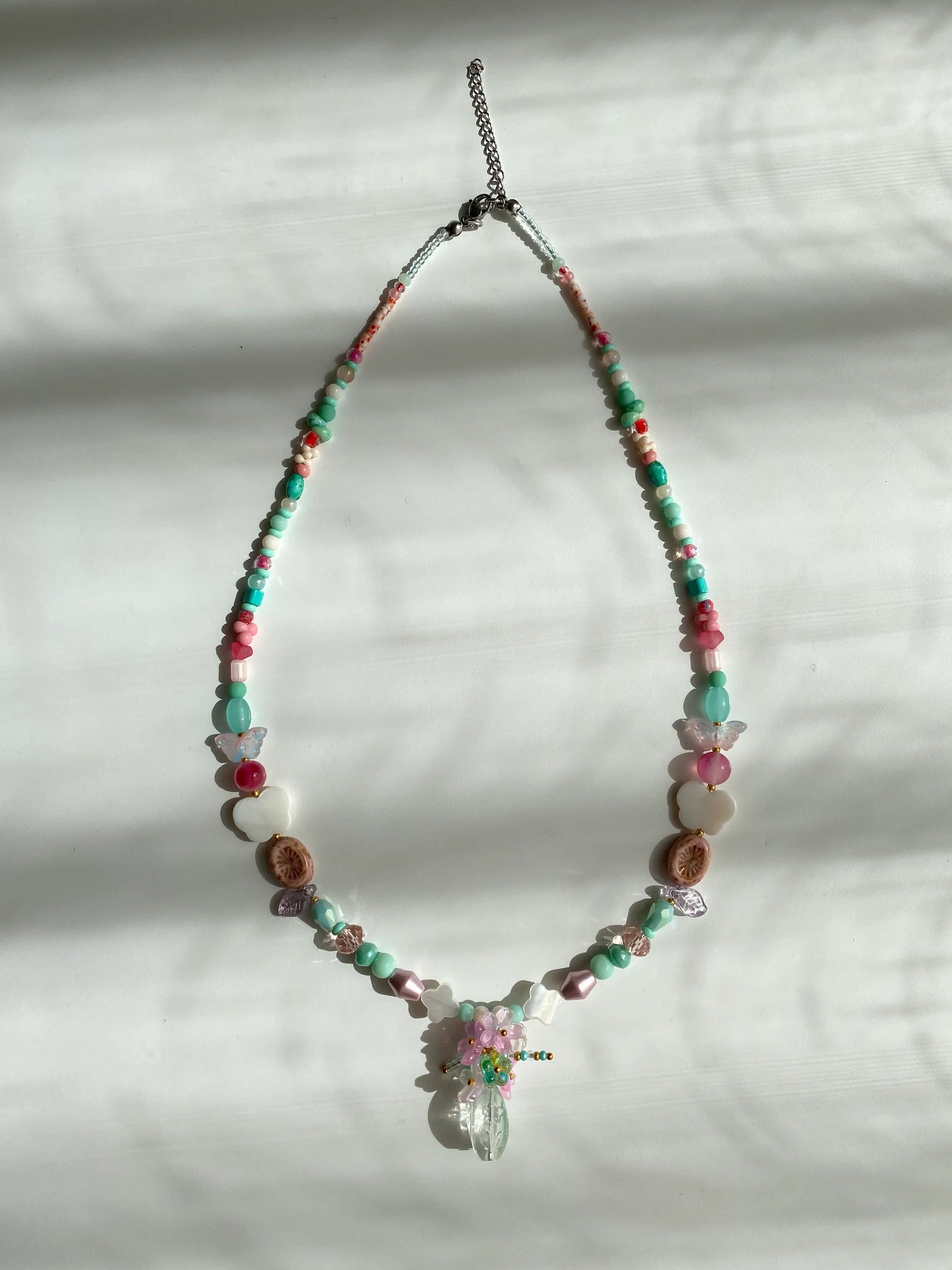 Detailed view of a unique pink and sky blue necklace with a beaded flower bouquet in a vase, featuring shell accents and kiwi glass beads on a sunlit white desk.