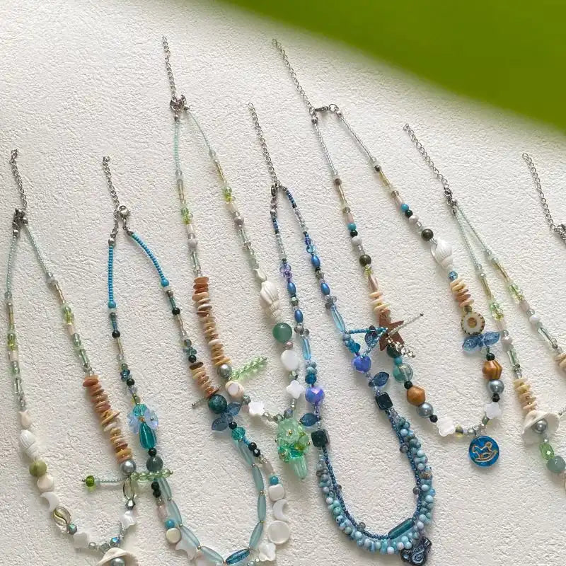 A vibrant flatlay of HuibenCraft handmade necklaces on a white table, featuring an ocean-inspired collection with blue-green beads, seashell charms, 3D floral accents, and intricate Czech glass beads.