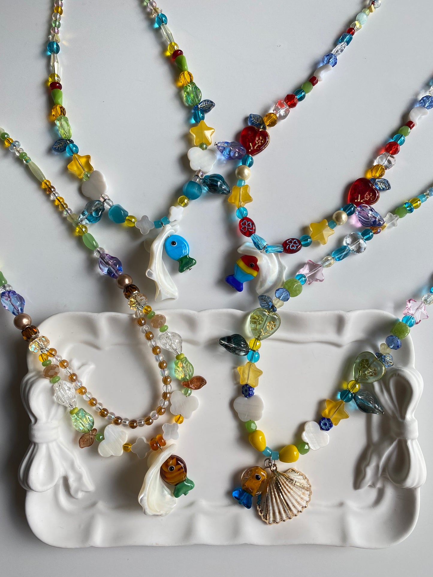 One-of-one handmade shell necklaces arranged on a ceramic plate with vibrant glass beads, butterfly shell charms and seaside-inspired details sparkling in the sunlight.