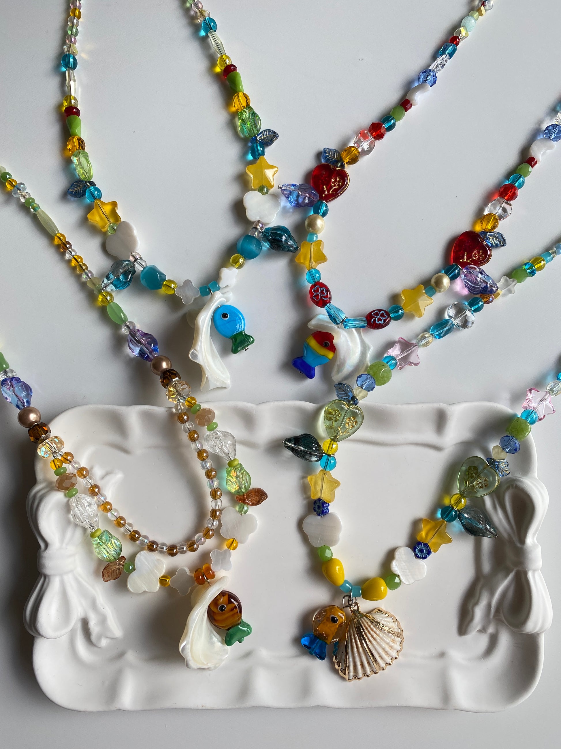 One-of-one handmade shell necklaces arranged on a ceramic plate with vibrant glass beads, butterfly shell charms and seaside-inspired details sparkling in the sunlight.