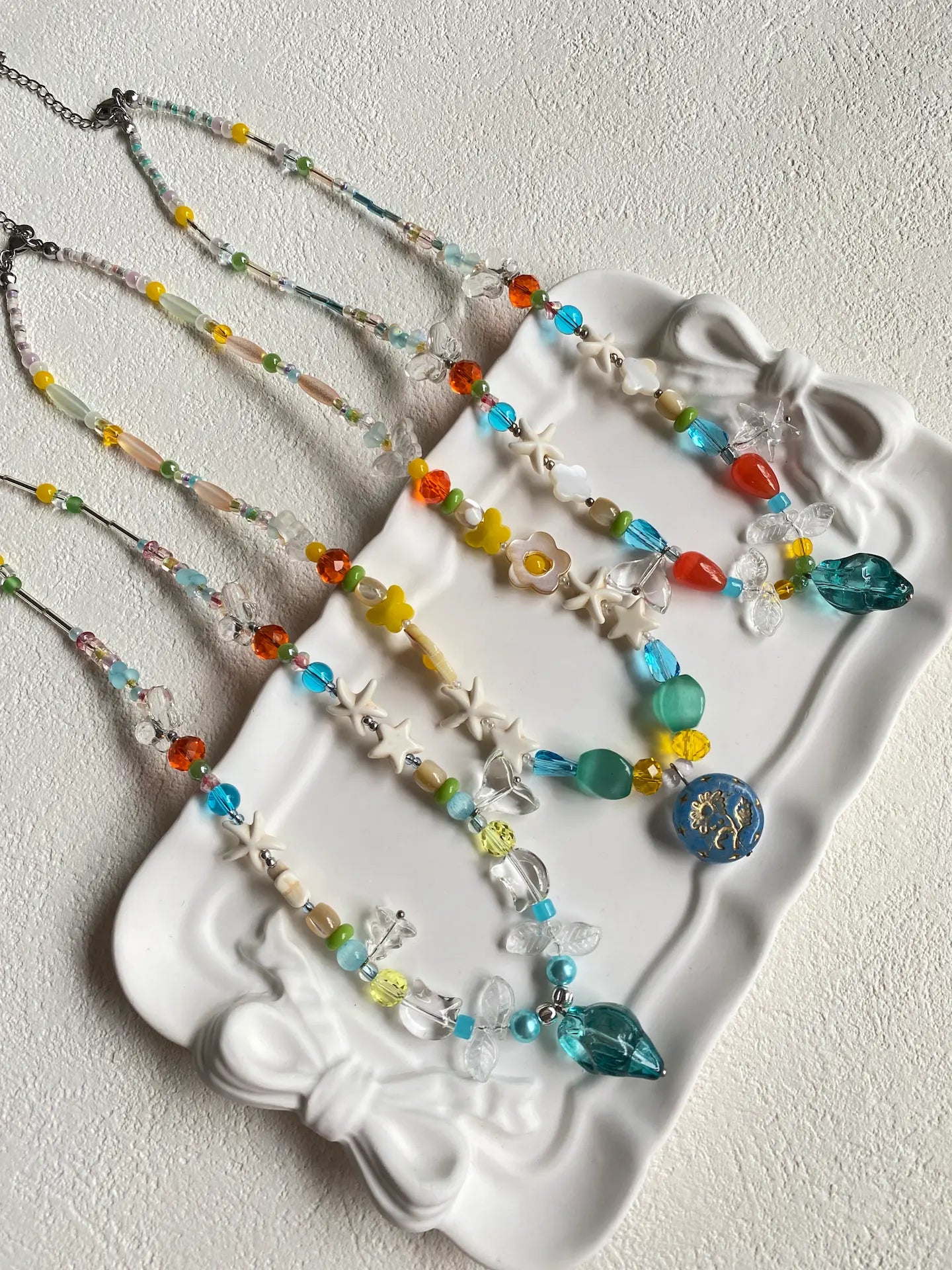 A trio of Huibencraft 1-of-1 ocean-themed necklaces featuring blue and green glass charms resting in a white dish.