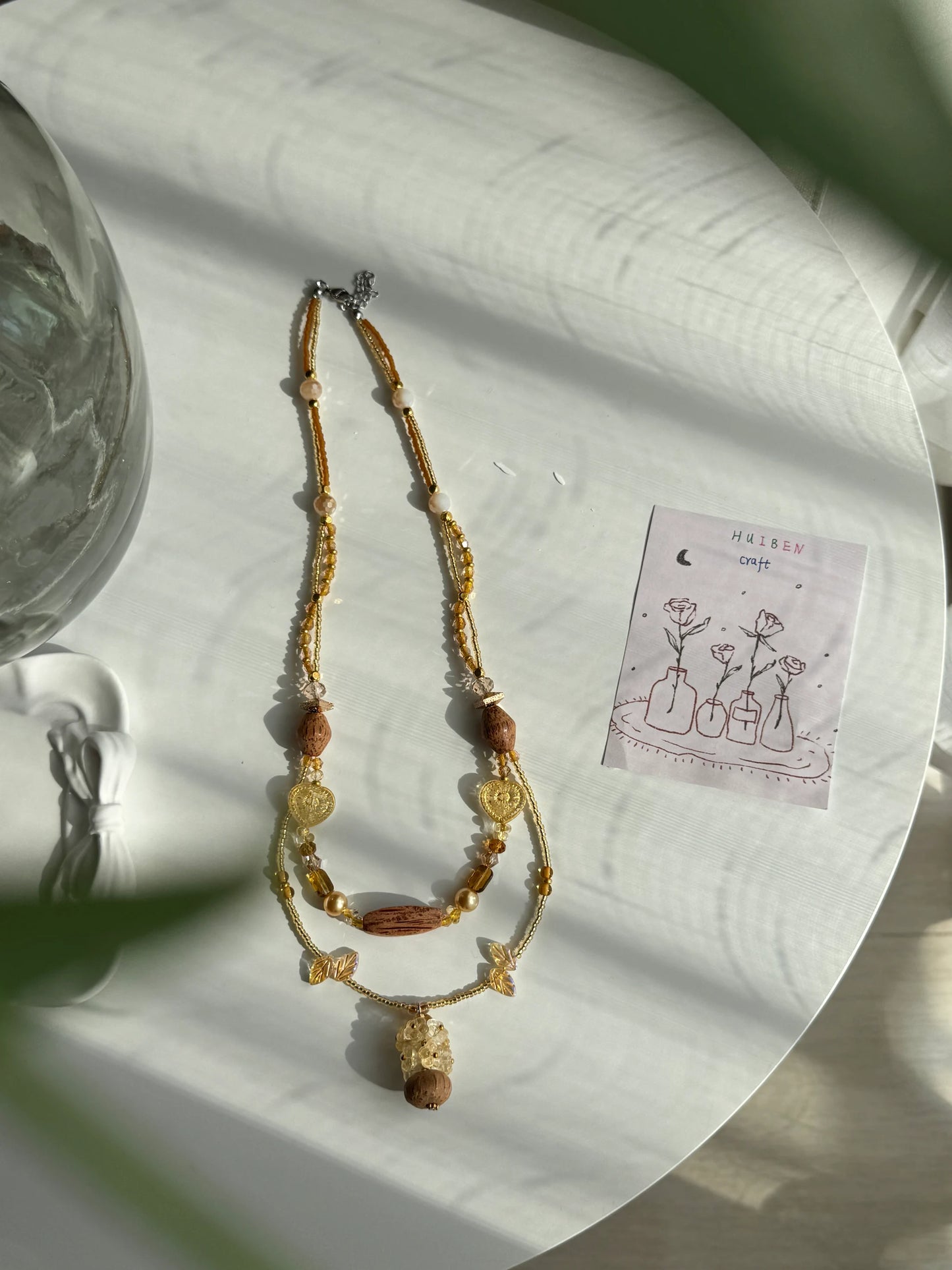Detailed view of a handcrafted khaki and coffee-colored beaded necklace with a voluminous 9-flower wooden vase charm on a sunlit white desk.