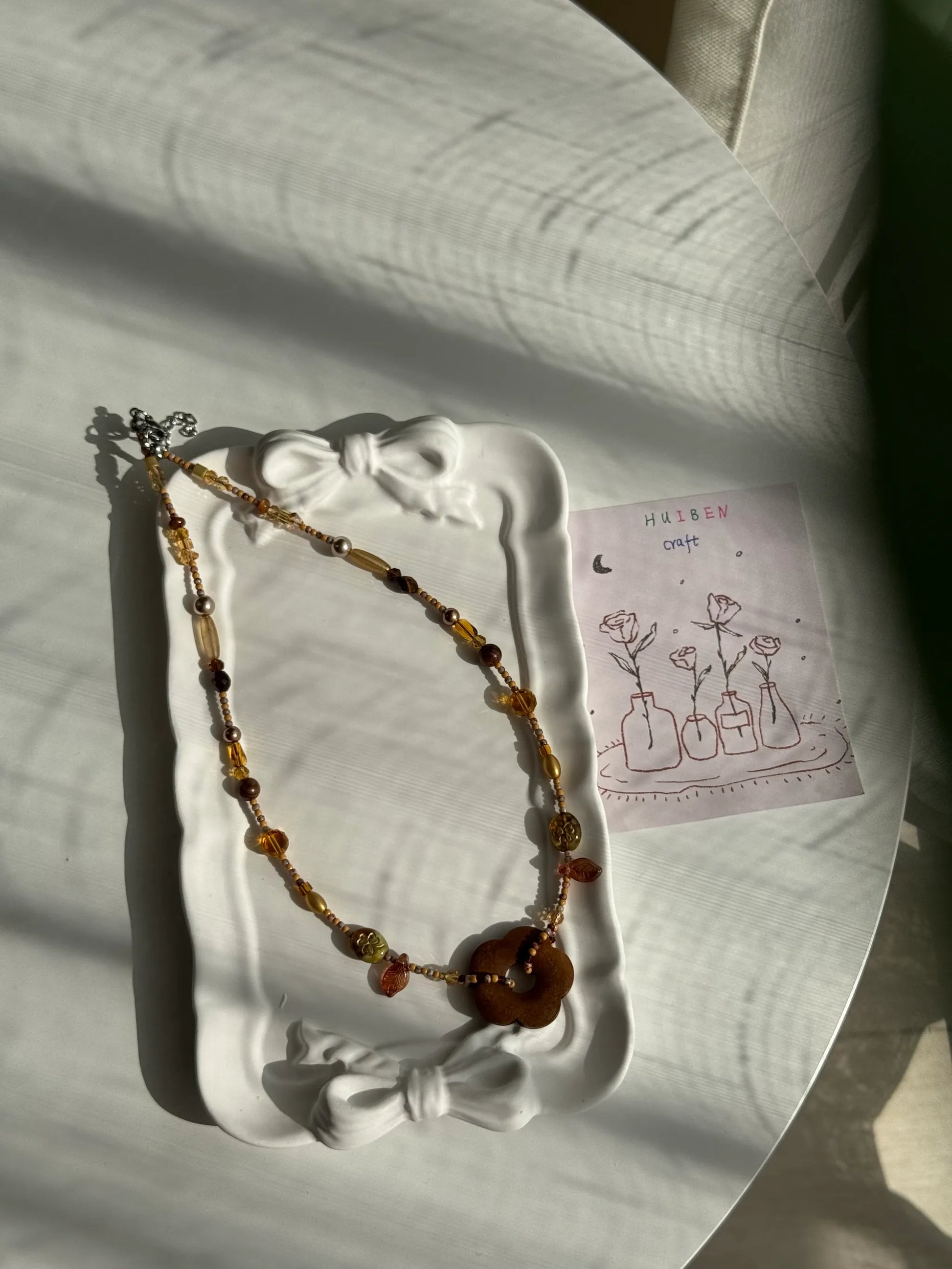 HuibenCraft 1-of-1 minimalist wooden flower beaded necklace with shimmering gold and coffee tones in a white ceramic dish.