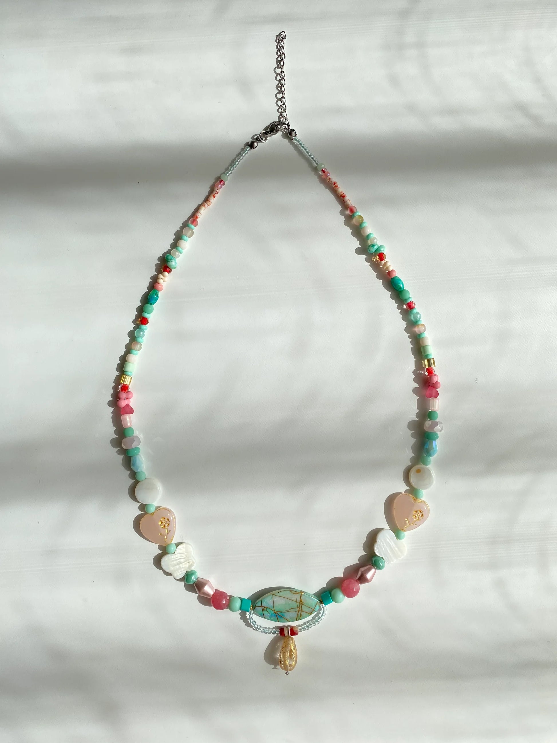 Detailed view of a handcrafted mixed-style necklace featuring shell butterflies, glass heart beads, and ethnic charms on a sunlit white desk.