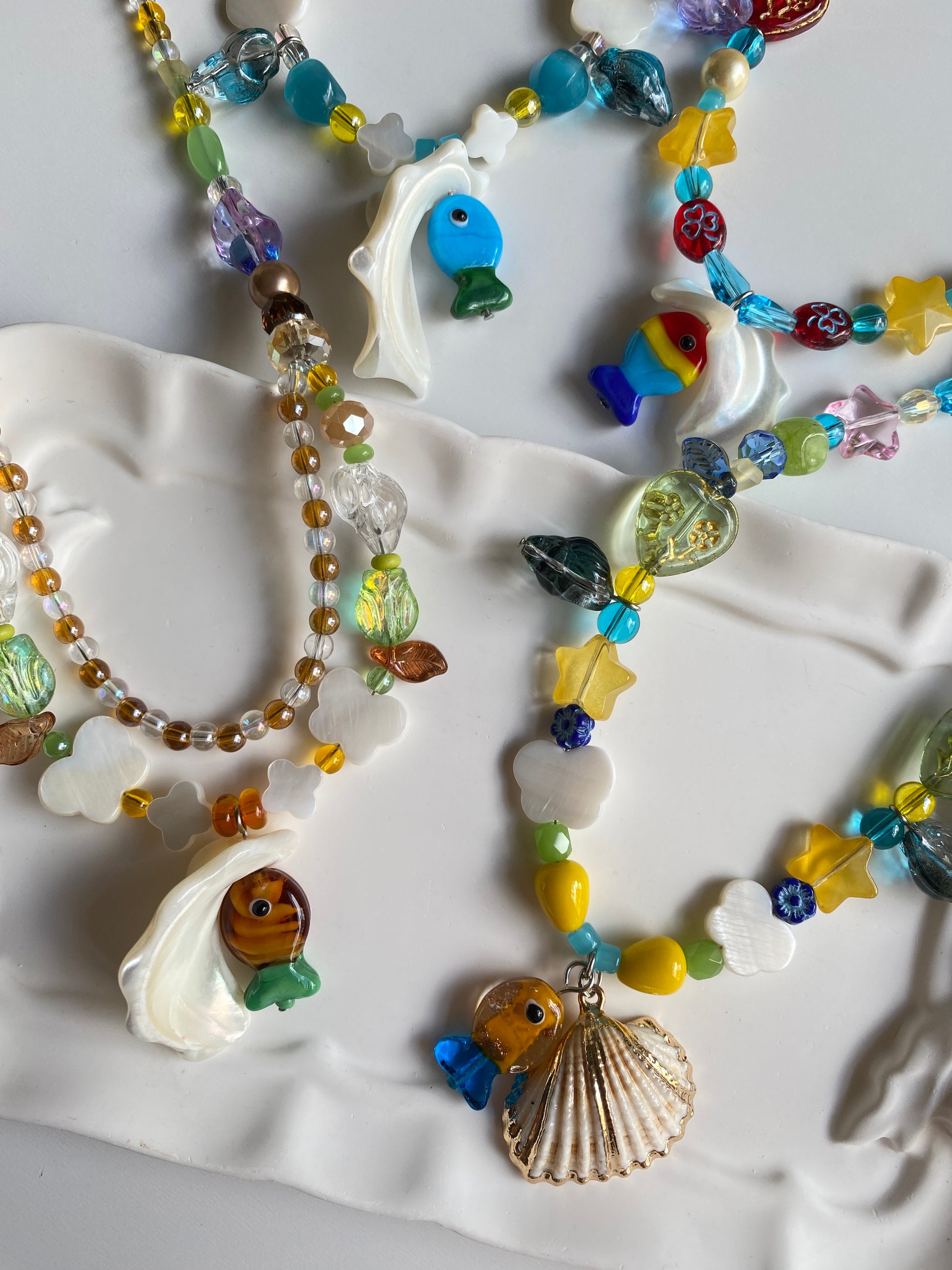Flat lay of four colorful handmade fish necklaces displayed on a white background, featuring natural shells, Czech glass fish, clover shell charms and summer-inspired beadwork.