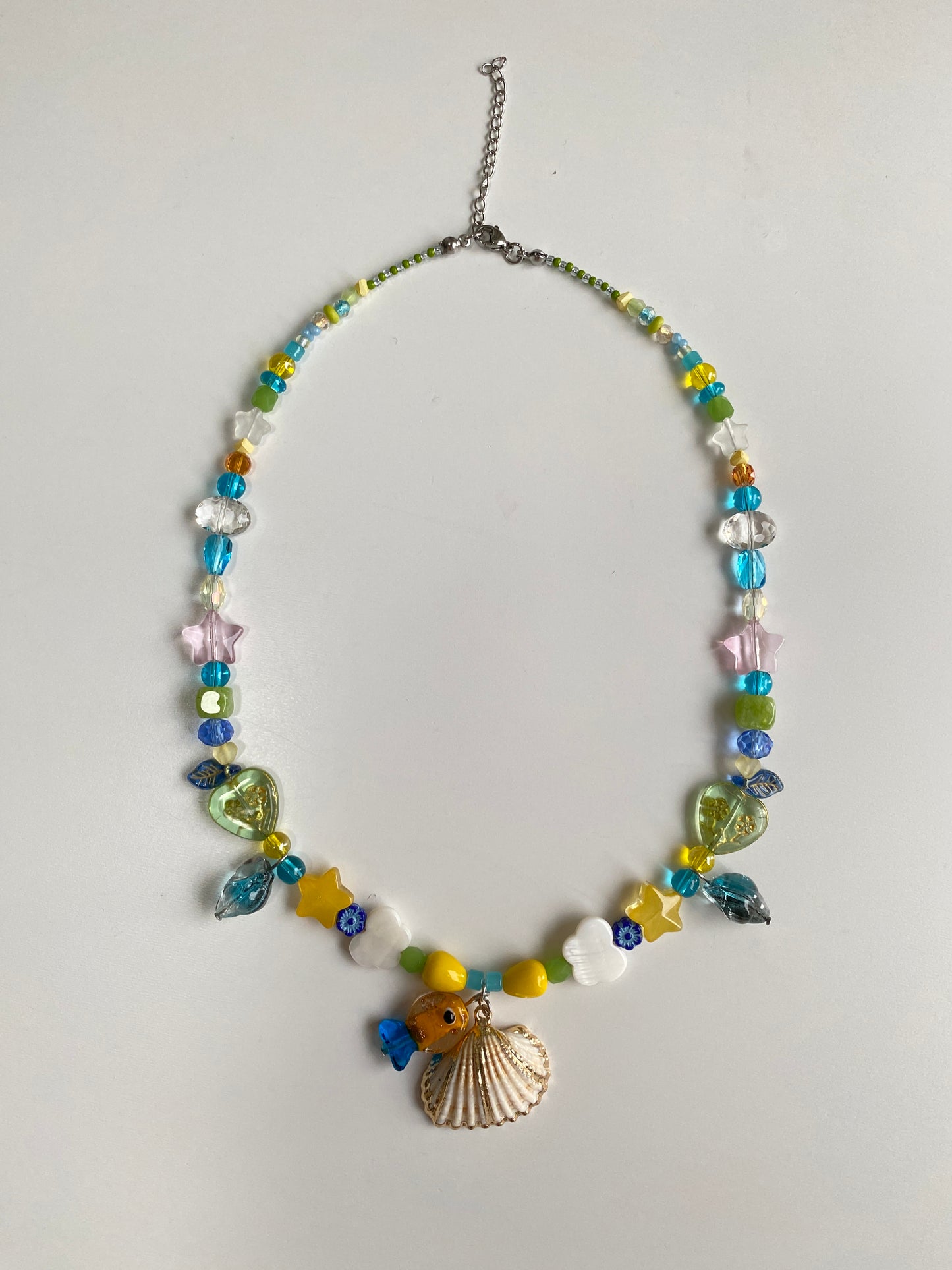 Close-up of a handmade ocean-inspired necklace featuring an orange-blue Czech glass fish, shell pendant, green heart glass bead and sparkling star details.