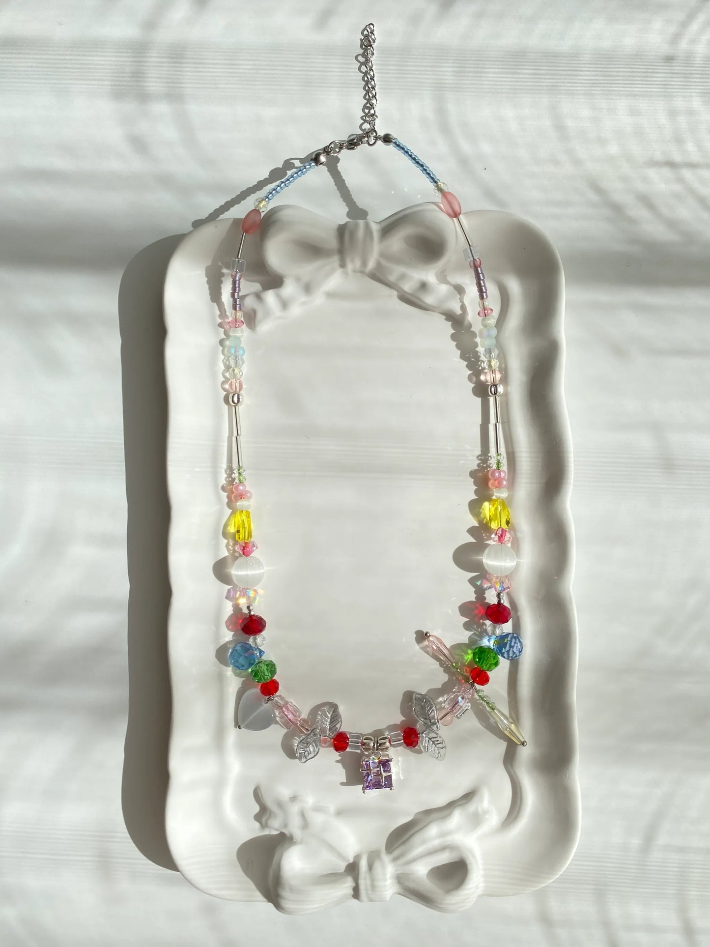 Unique handmade multi-color seed bead rainbow necklace with side dangle details and a purple pendant, displayed beautifully in a small dish.