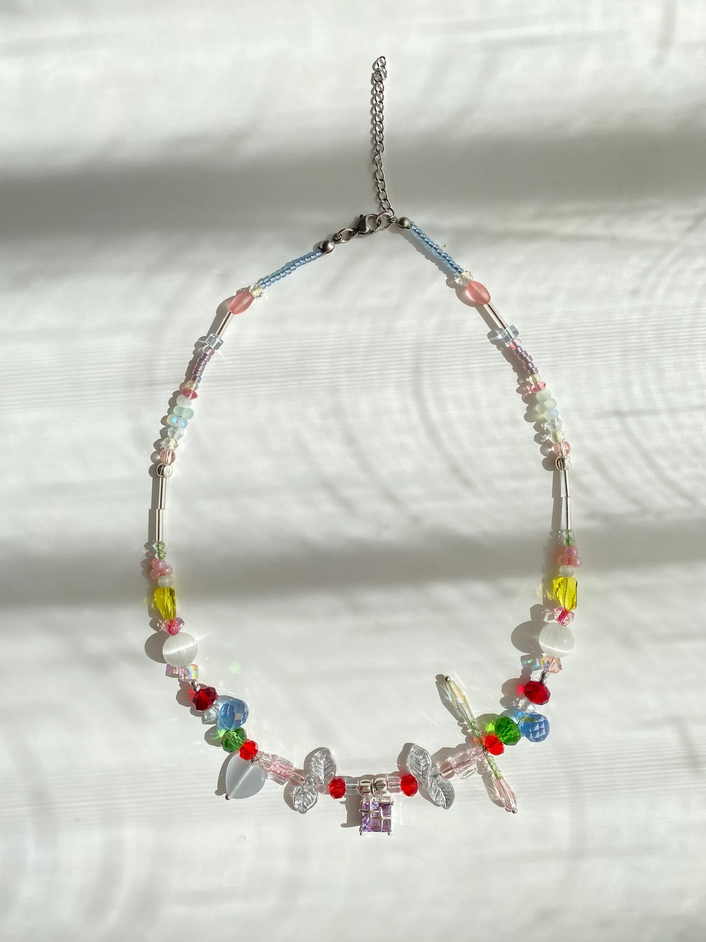 Vibrant rainbow beaded choker featuring leaf and teardrop accents with a purple glass pendant, flat lay on a table in sparkling sunlight.