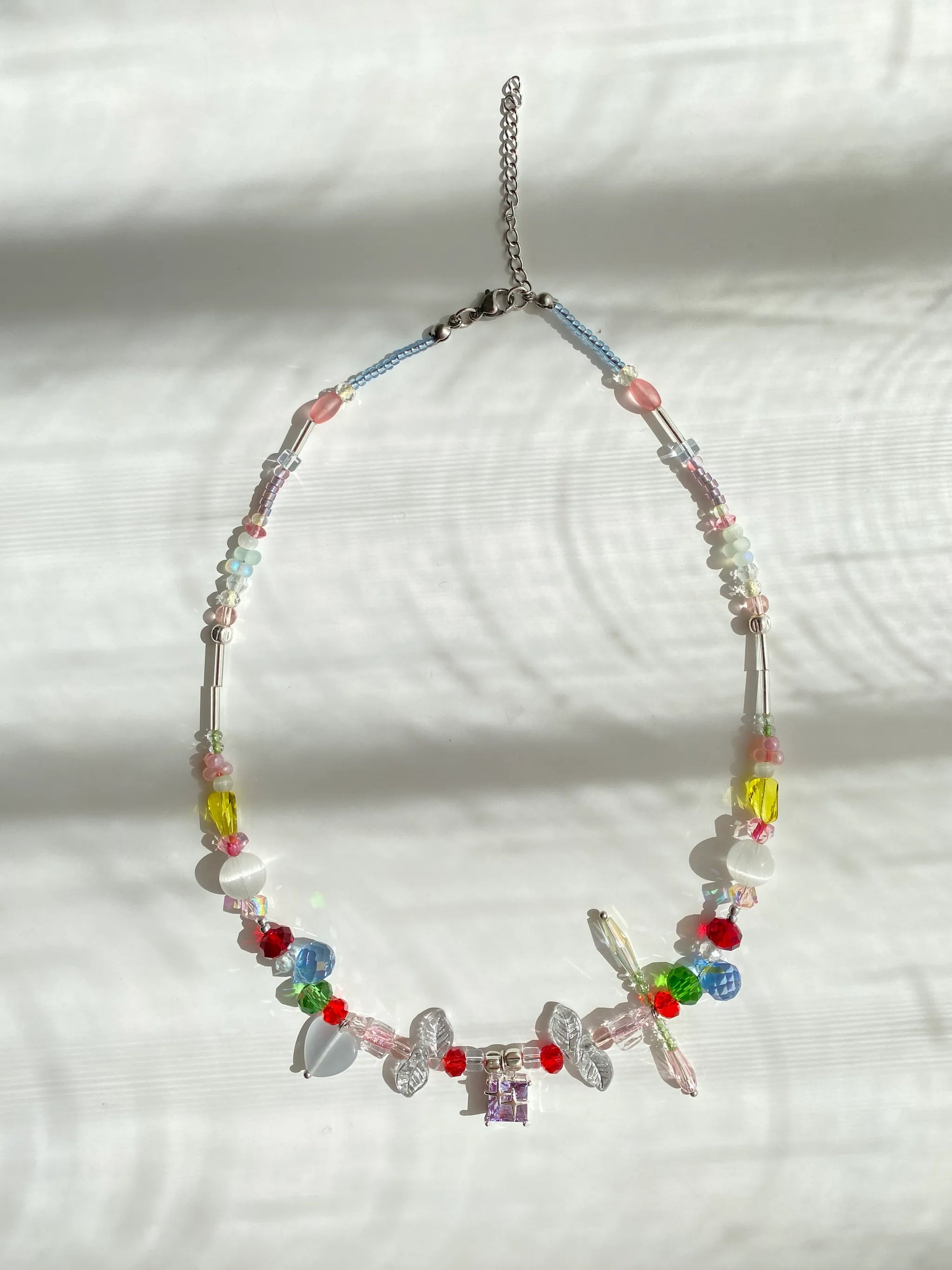 Vibrant rainbow beaded choker featuring leaf and teardrop accents with a purple glass pendant, flat lay on a table in sparkling sunlight.