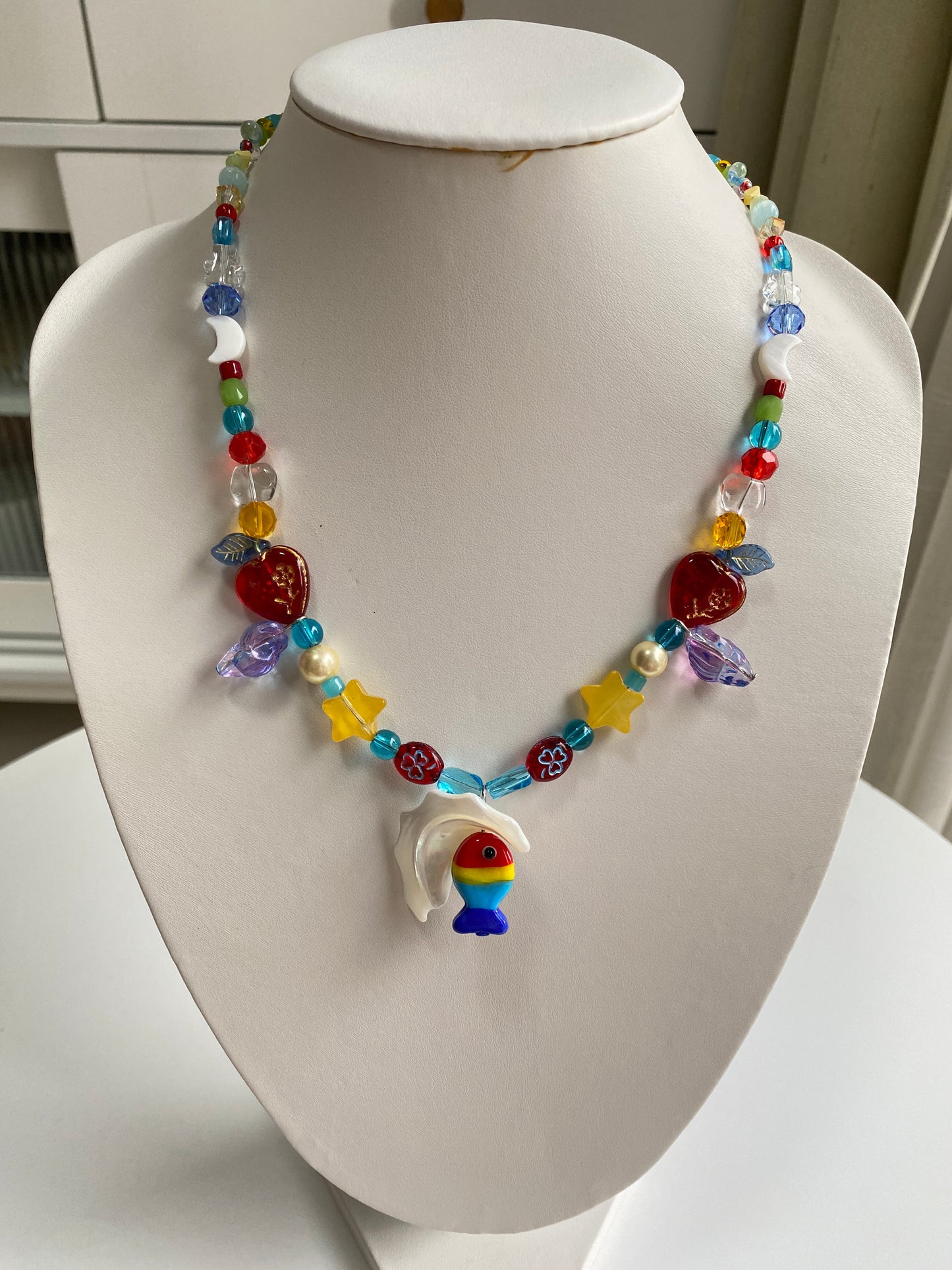 Close-up of a handmade fish necklace featuring a red, yellow and blue glass fish, star bead, moon shell and gold-trimmed heart glass accents.