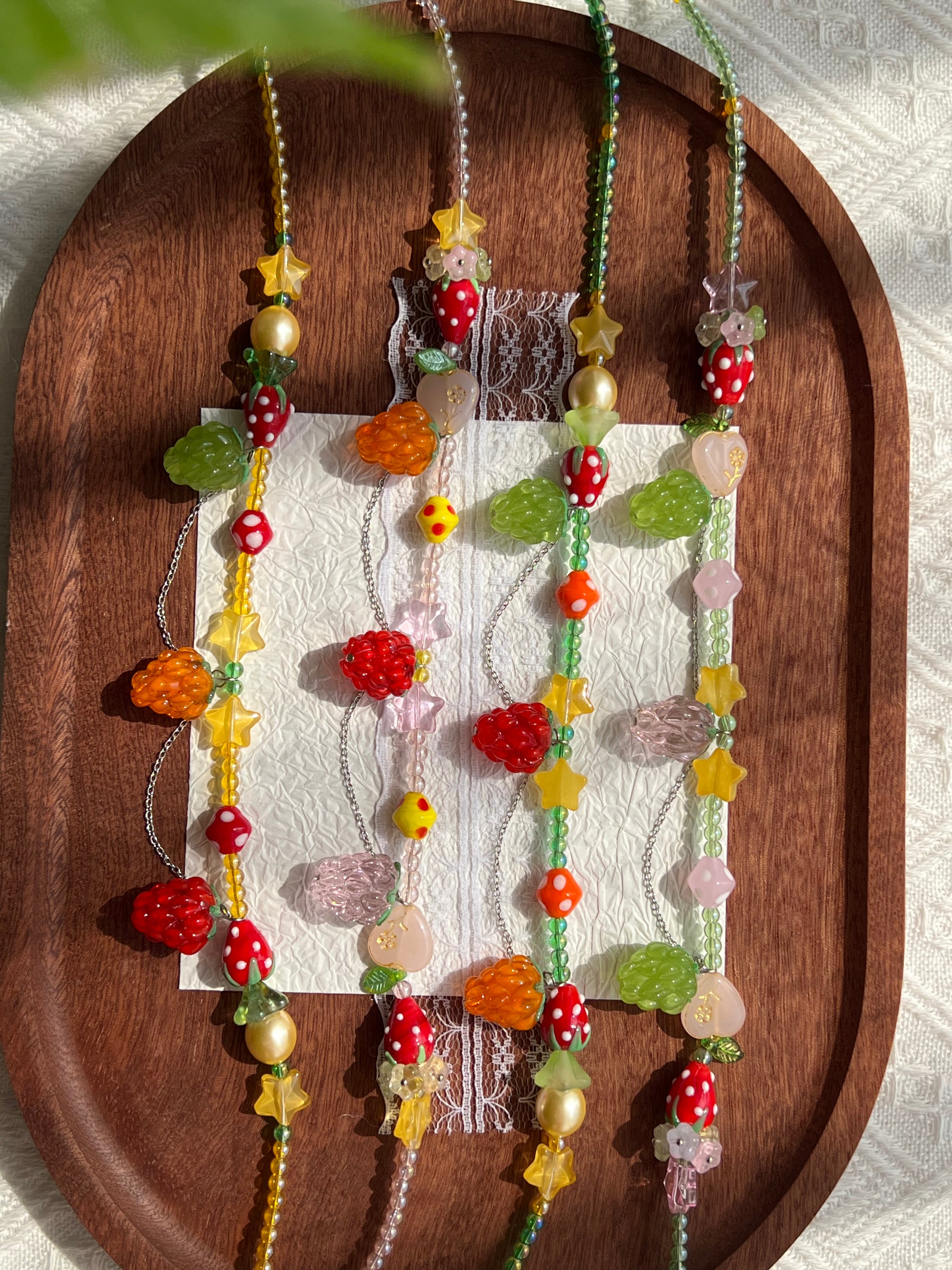 Overhead view of four raspberry and strawberry glass bead necklaces arranged on a wooden tray with white paper and lace.