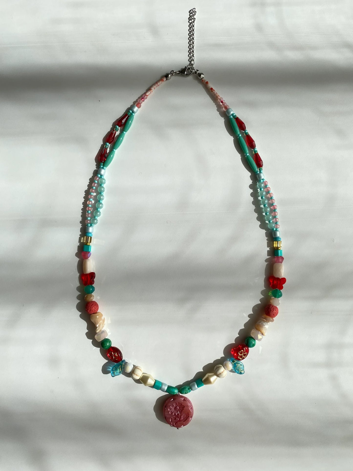 Detailed view of a handcrafted necklace featuring red glass butterflies, clovers, white pine stones, and shells on a sunlit white desk.
