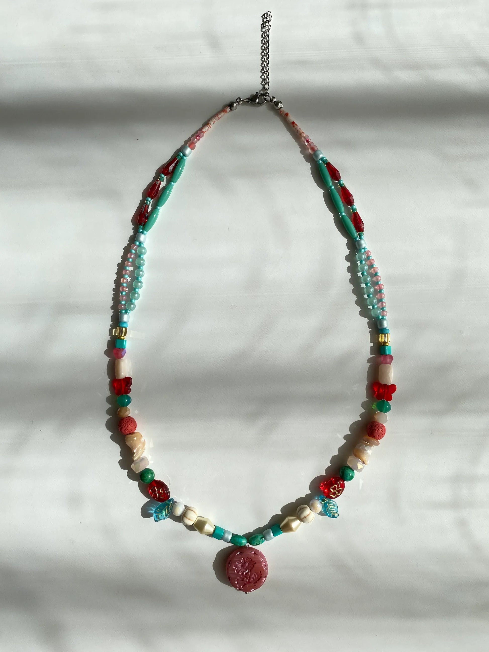 Detailed view of a handcrafted necklace featuring red glass butterflies, clovers, white pine stones, and shells on a sunlit white desk.