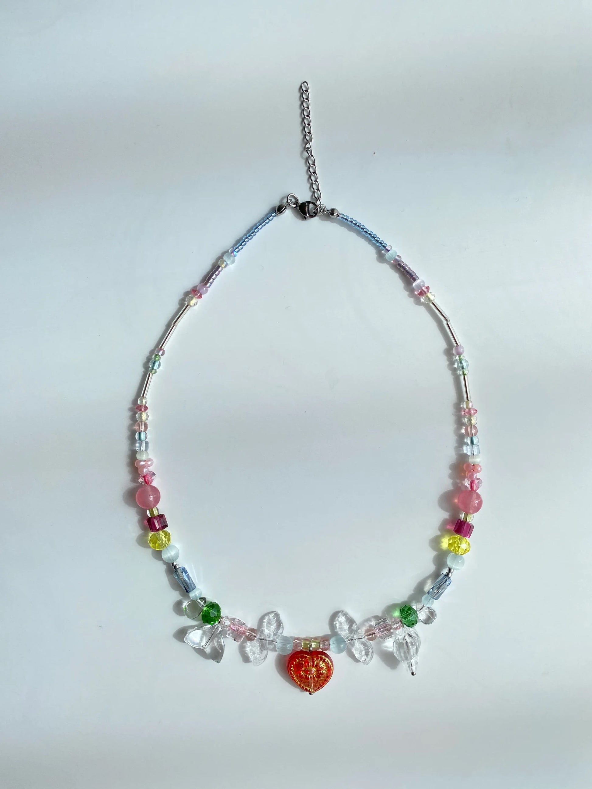 Artisan handmade colorful choker featuring a red heart glass pendant, clear conch shells, and pink stones, flat lay on a table in brilliant sunshine.