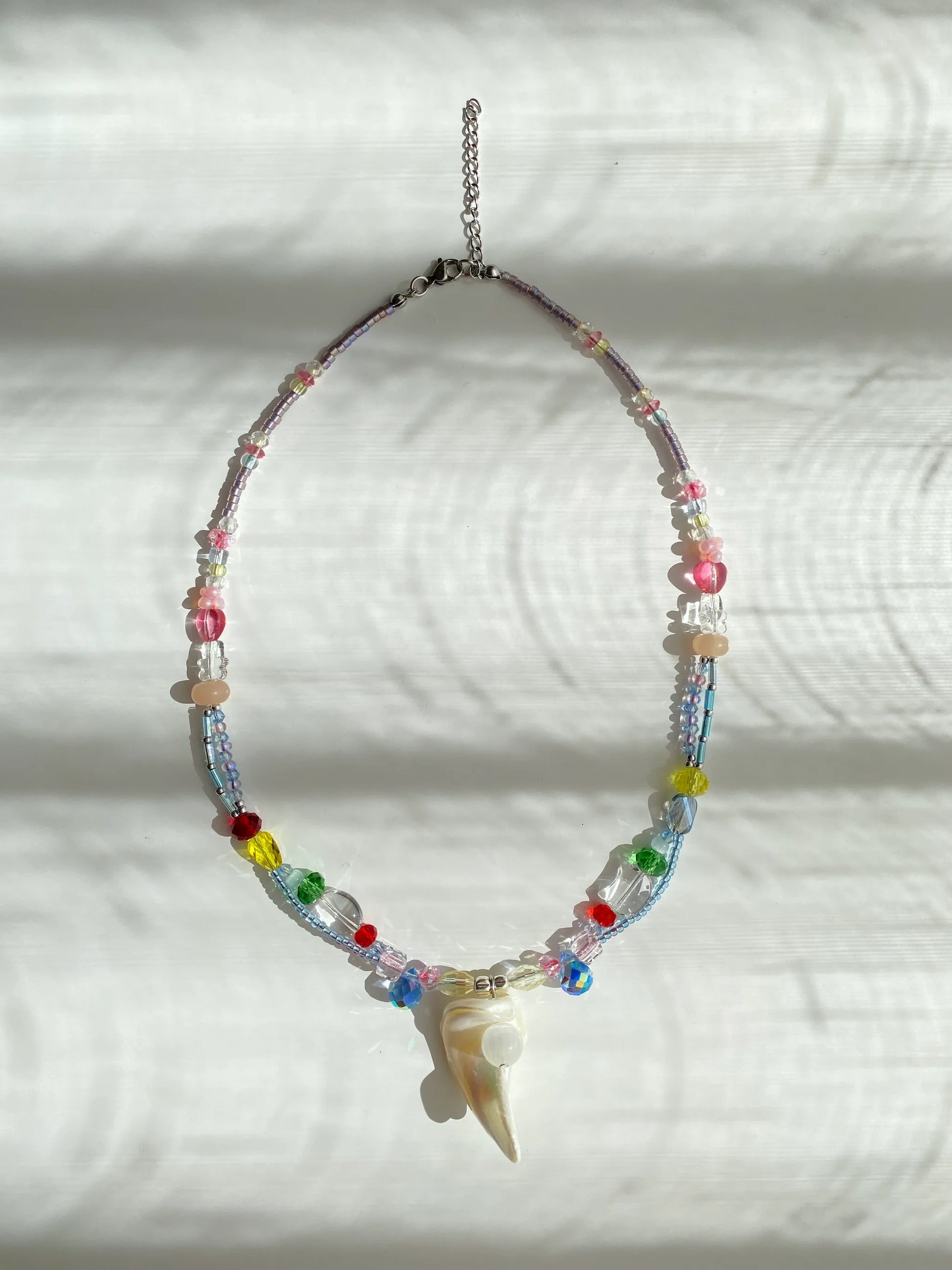 Artisan handmade multi-color seed bead necklace with shell and cat's eye bead pendant, flat lay on a table in sun-drenched lighting.