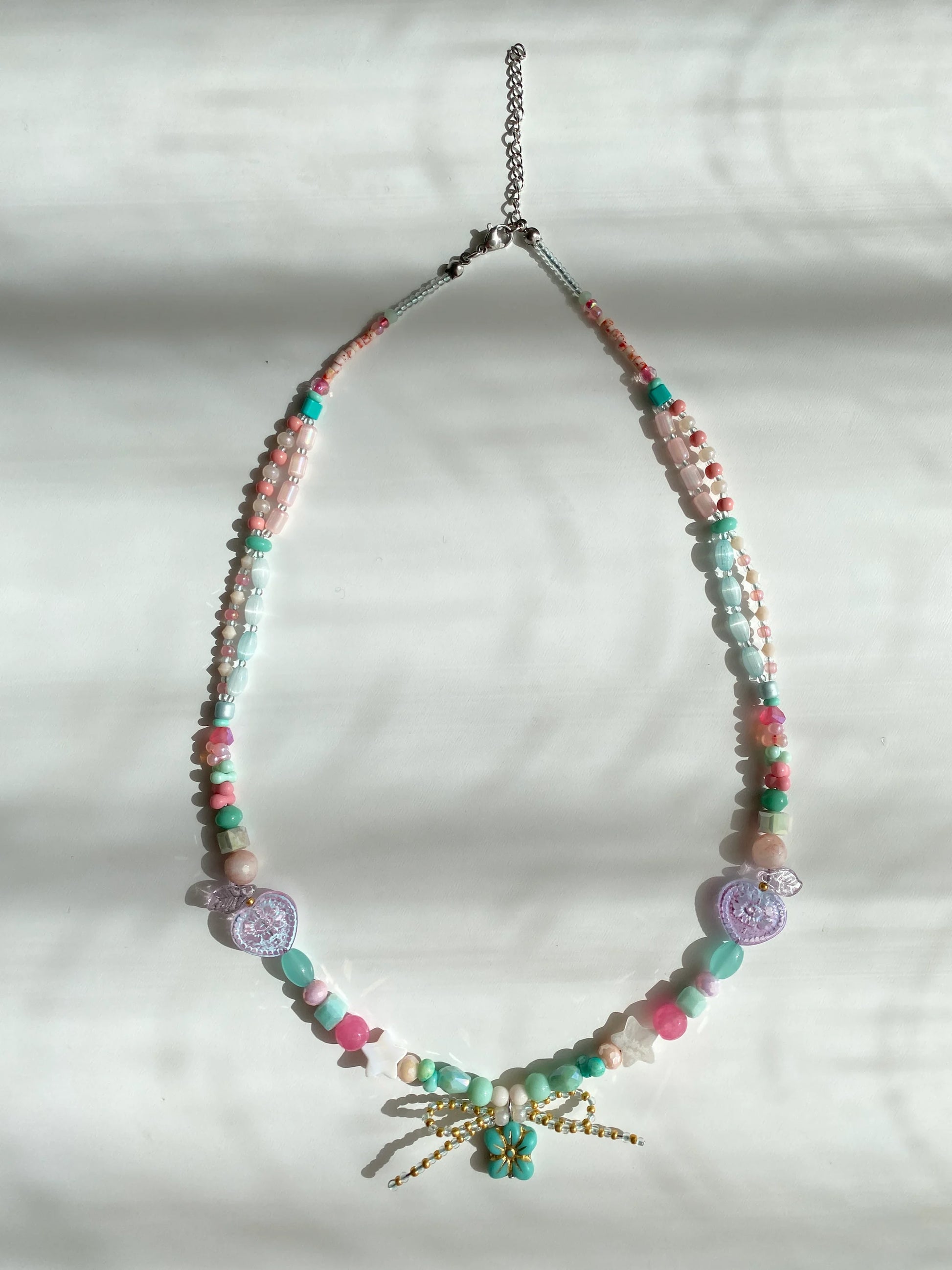 Detailed view of a colorful handcrafted necklace featuring shell stars, glass hearts, and a dainty bead bow on a sunlit white desk.