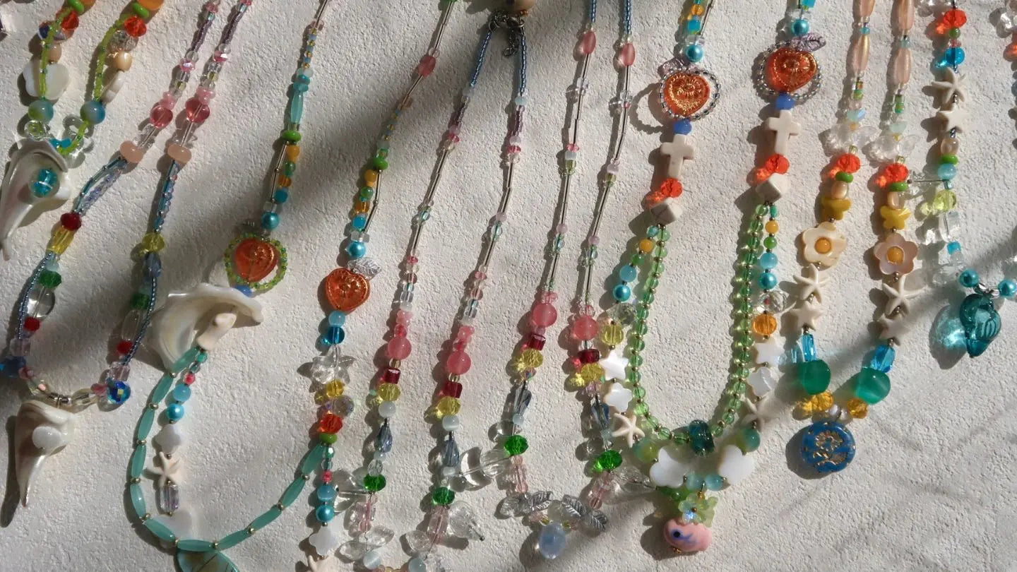 A vibrant, joyful collection of HuibenCraft 1-of-1 summer beaded necklaces on a clean white table, featuring colorful transparent beads, playful glass ducks, and seashell charms for a whimsical ocean aesthetic.
