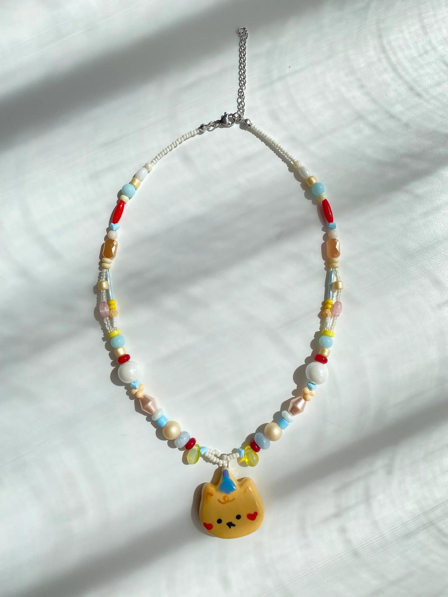 Flatlay of a handmade pastel beaded necklace with a proud bear pendant on a white table.