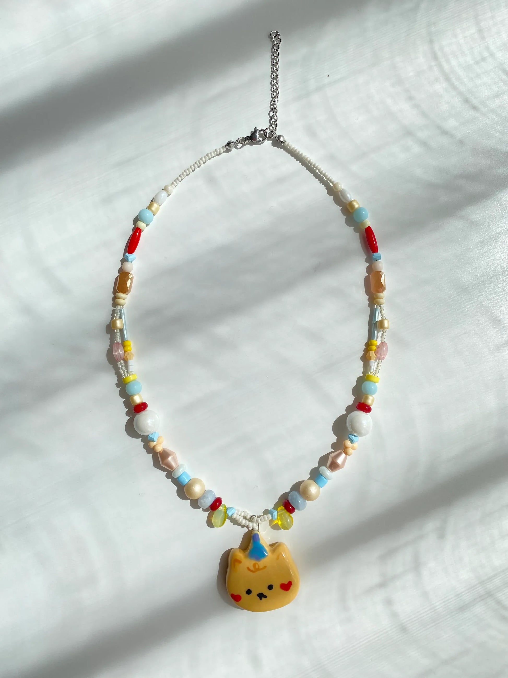 Flatlay of a handmade pastel beaded necklace with a proud bear pendant on a white table.