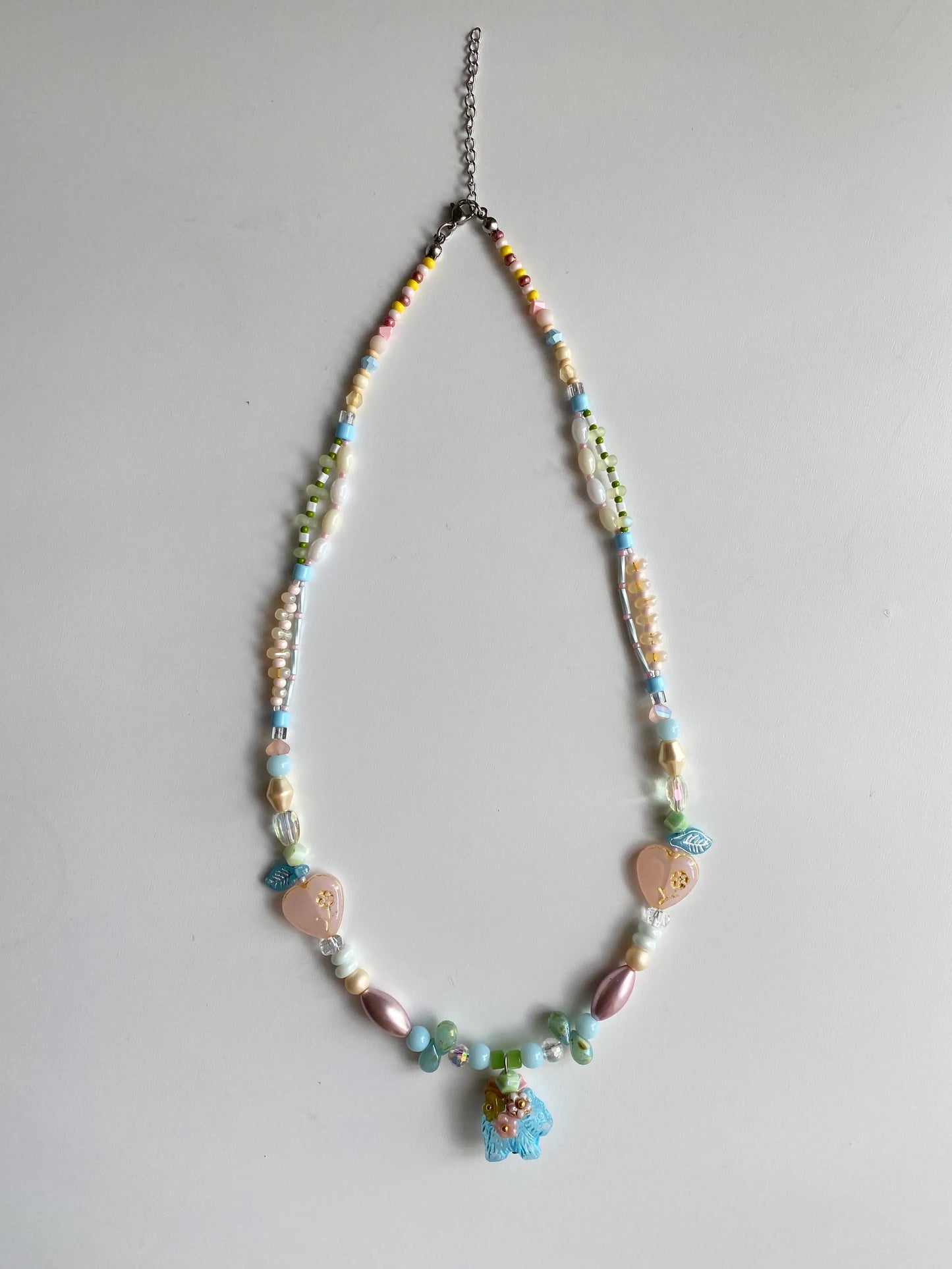 Flatlay of a handmade pastel beaded necklace featuring heart and dog charms on a white table.