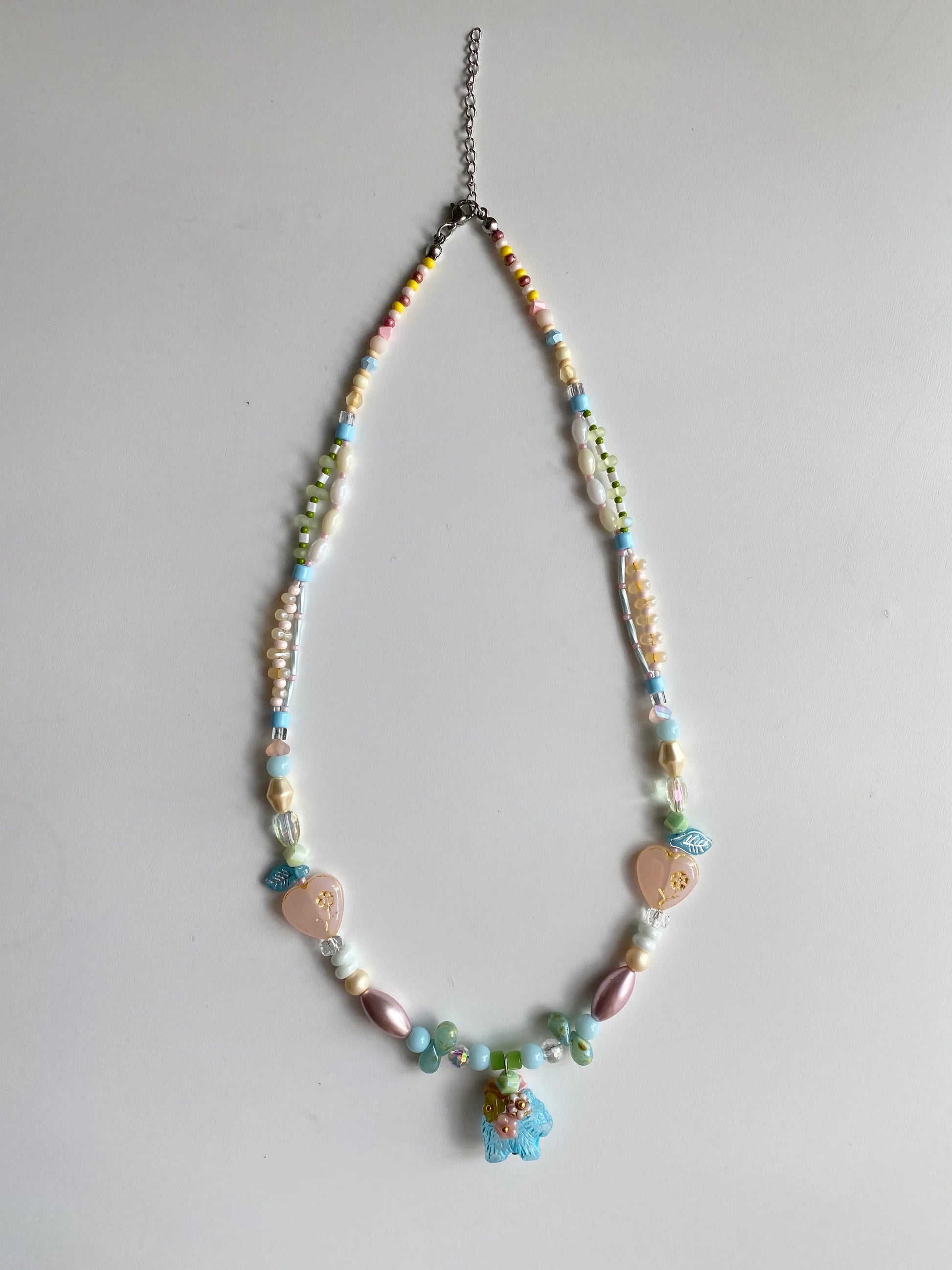 Flatlay of a handmade pastel beaded necklace featuring heart and dog charms on a white table.